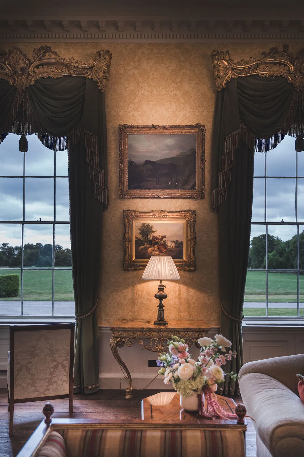 A lamp between two tall sash windows hung with paintings