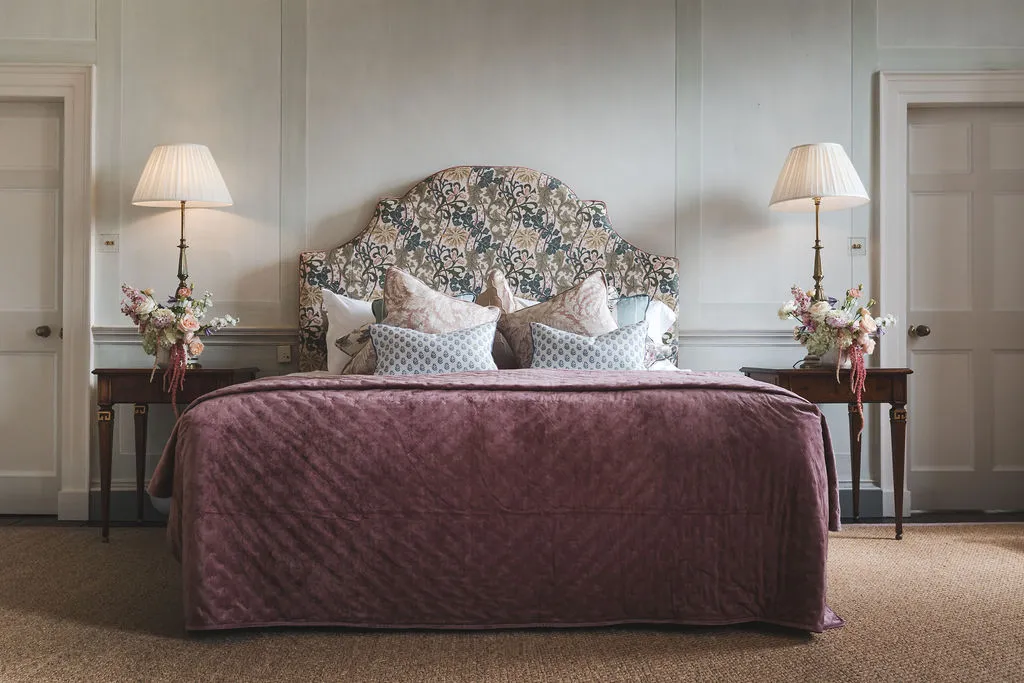 An upholstered headboard and velvet quilt in a Manor House suite, flowers either side